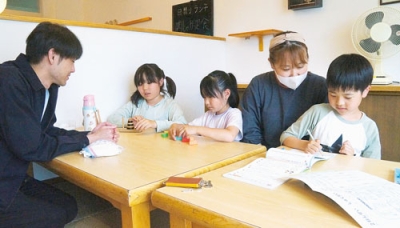 写真：田村店長（右から２人目）、佐野さん（左）に手伝ってもらいながら宿題や立体パズルに熱中する児童たち