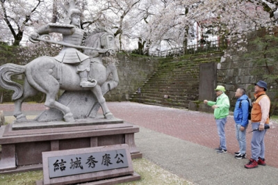 写真：福井城を築いた結城秀康の像の前で、観光客に説明する語り部（左）＝４月、福井市