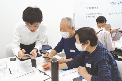 写真：ボランティア学生（左）の助言を受け、電話操作を学ぶ受講者