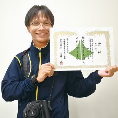 写真：愛用するカメラと賞状を手に入賞を喜ぶ田中さん