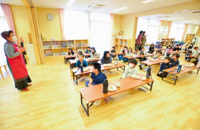 写真：對馬理事長（左）から料理やかえる食堂について説明を受ける児童ら