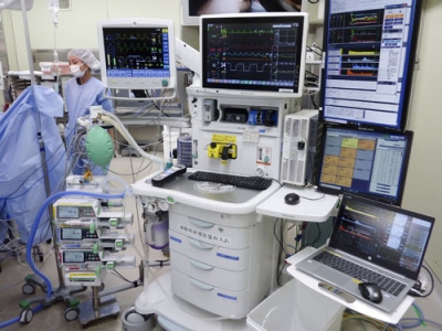 写真：稼働中のロボット麻酔システム。右手前がソフトウエアを組み込んだパソコン、左手前が３種類の麻酔薬を注入するシリンジポンプ。左奥で全身麻酔の手術が進行中＝福井県永平寺町の福井大病院